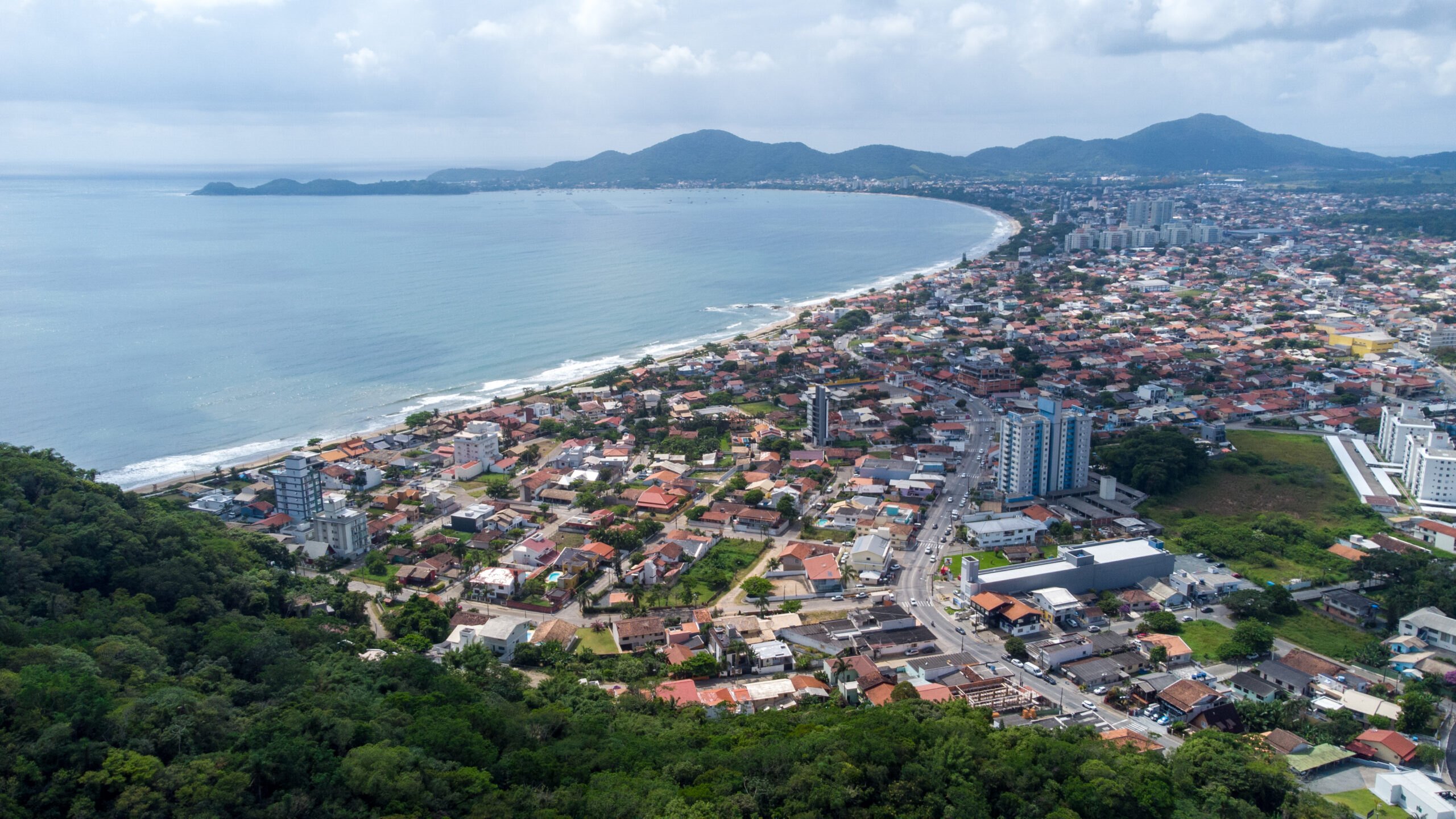 Vista aérea da orla de Penha, com a enseada, a faixa de praia e a cidade ao fundo das montanhas