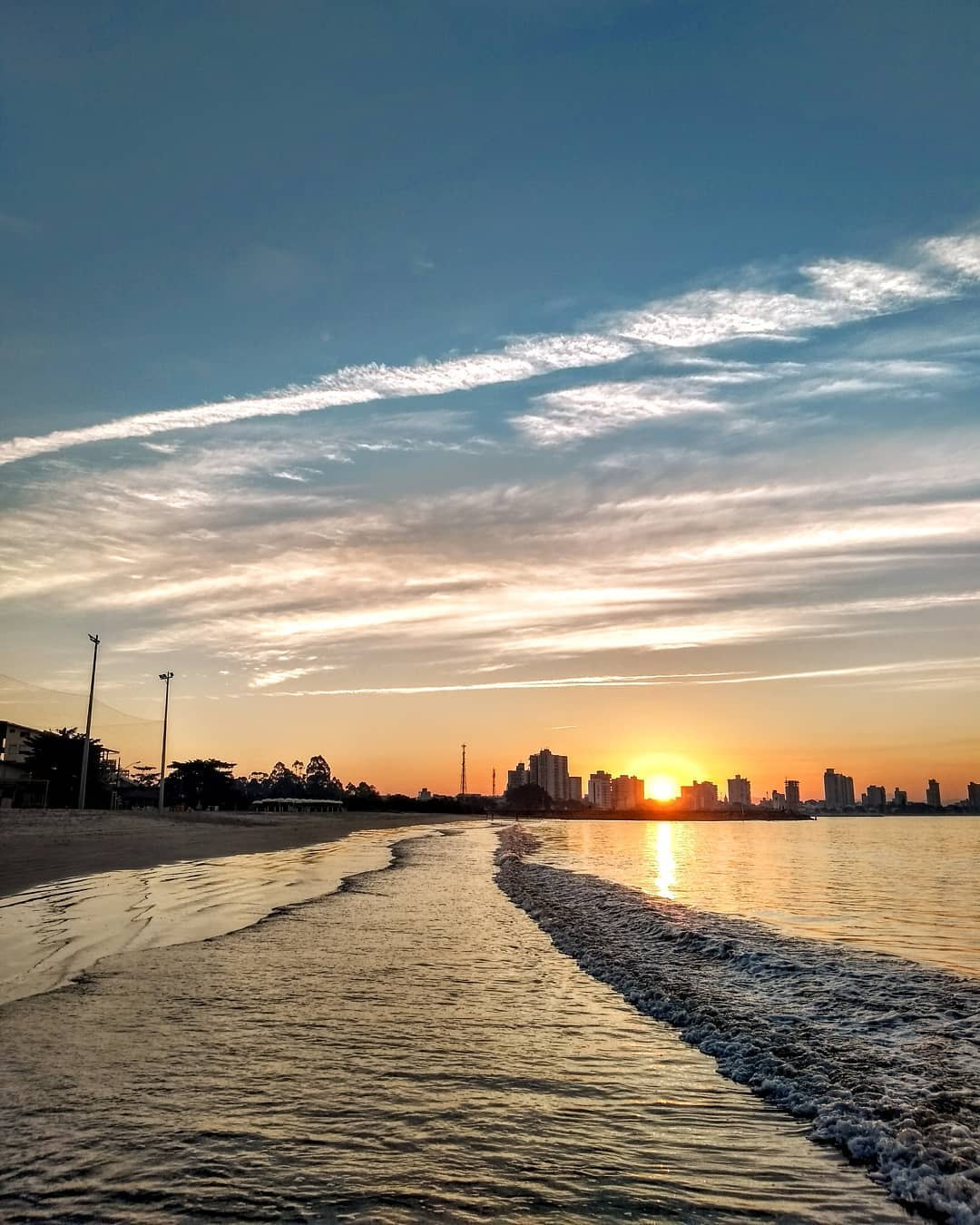 Pôr do sol na Praia Alegre, em Penha, com o skyline da cidade ao fundo refletido na água